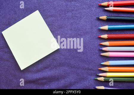 Mehrfarbige, leuchtende, farbenfrohe Bleistifte zum Zeichnen befinden sich auf der rechten Seite und ein Notizbuch auf der linken Seite für Ihren Text auf einem lilafarbenen Hintergrund. Stockfoto