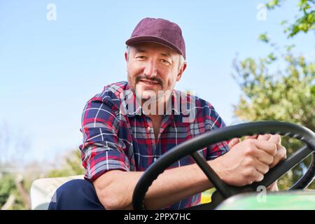 Porträt eines reifen männlichen Landwirts, der am sonnigen Tag einen Traktor mit klarem Himmel im Hintergrund fährt Stockfoto