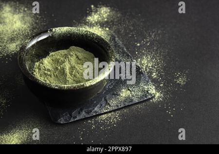 Grünes Pulver in einer kleinen Keramikschüssel, auf einem Steinständer, auf schwarzem Hintergrund. Der Hintergrund ist mit diesem Pulver bestreut. Super Food oder ma Stockfoto