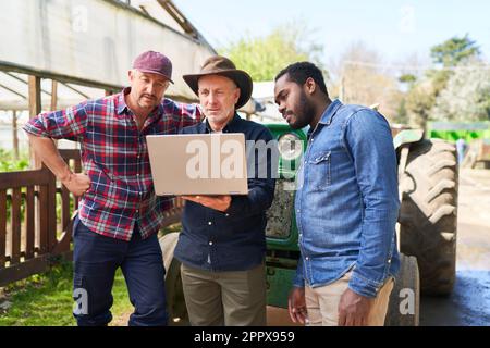 Reifer männlicher Landwirt, der mit Kollegen über einen Laptop vor dem Traktor auf dem Bauernhof spricht Stockfoto