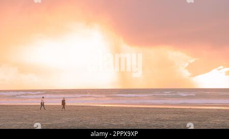 Zwei Personen, die bei Sonnenuntergang an einem Sandstrand spazieren, Neuseeland Piha bei Sonnenuntergang Stockfoto