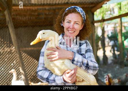 Porträt einer glücklichen Farmerin mit lockigem Haar und weißer Gans auf dem Bauernhof Stockfoto