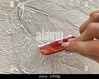 Die Künstlerin hält einen Kamm in der Hand und trägt eine Textur aus volumetrischer Acrylfarbe auf die Leinwand des Gemäldes auf. Stockfoto