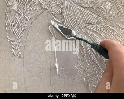 Die Hand des Künstlers besputzt die Leinwand mit einem kleinen Spatel mit volumetrischer Acrylfarbe in der kreativen Kunst-Werkstatt des Künstlers mit Farben und Stockfoto