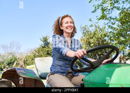 Glückliche, reife Landwirtin, die an sonnigen Tagen auf einem grünen Traktor mit klarem Himmel im Hintergrund sitzt Stockfoto