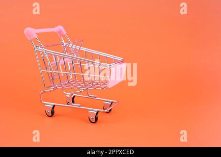 Kleiner Supermarkt, Einkaufswagen mit isolierten Rädern auf orangefarbenem, pastellfarbenem, trendigen Papierhintergrund. Stockfoto