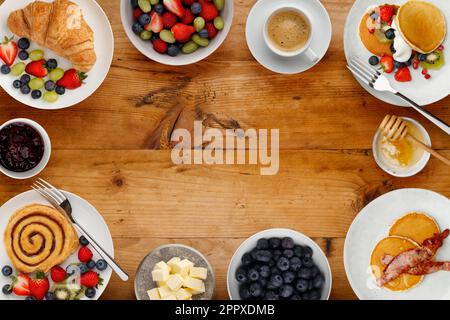 Ein Rand mit köstlichen Frühstückstellern, Pfannkuchen und Gebäck, Schalen mit Obst und Haferbrei und Tassen Kaffee auf rustikalem Holzhintergrund Stockfoto