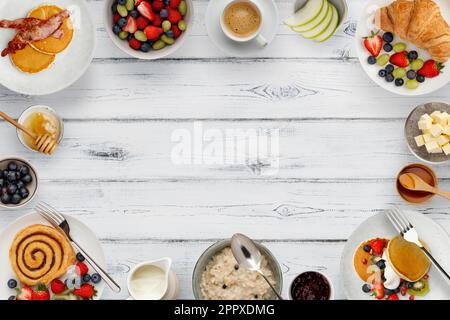 Ein Rand mit köstlichen Frühstückstellern, Pfannkuchen und Gebäck, Schüsseln mit Obst und Haferbrei und Tassen Kaffee auf rustikalem weißem Holzhintergrund Stockfoto