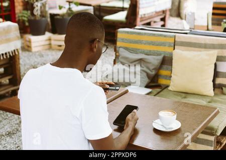 afroamerikanischer Mann sitzt am Tisch im Café mit einer Tasse Kaffee auf der Sommerveranda, hält ein Smartphone und gibt die Kreditkartennummer ein. Rückansicht Stockfoto