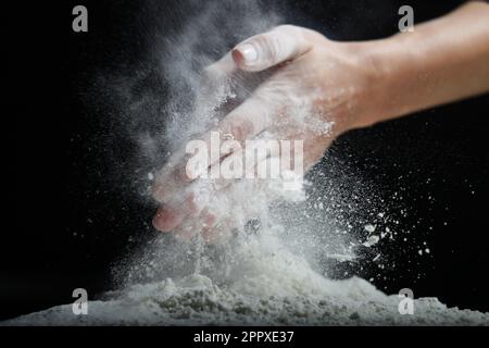 Weibliche Hände sieben Mehl mit einem Metallsieb auf schwarzem Hintergrund. Stockfoto