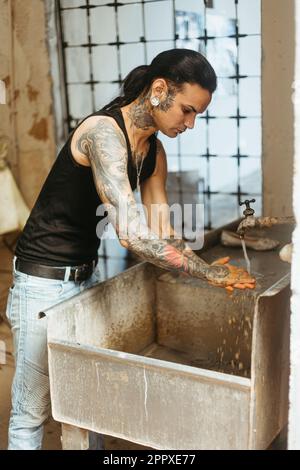Seriöser junger tätowierter männlicher Meister in legerer Kleidung mit langen Haaren, der schmutzige Hände aus Lehm wäscht, in einer professionellen Töpferwerkstatt Stockfoto