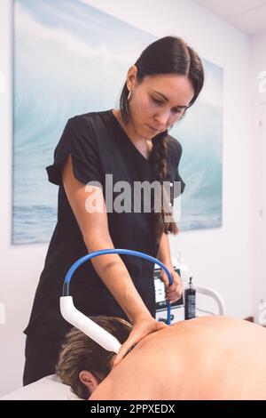 Junge Therapeutin in Uniform, die nach unten schaut und auf dem Rücken eines nicht erkennbaren Patienten arbeitet, der auf dem Bauch auf dem Tisch liegt und in der Klinik mit Geräten befestigt ist Stockfoto