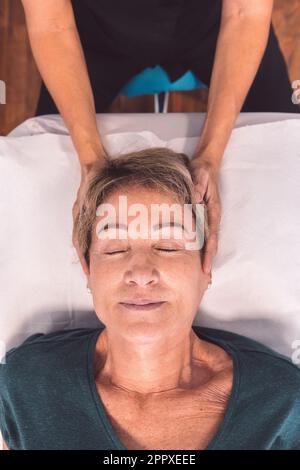 Von oben eine nicht erkennbare Therapeutin, die langsame Bewegungen mit den Fingern an den Kopf einer Seniorin anlegt, die mit geschlossenen Augen in der Klinik liegt Stockfoto