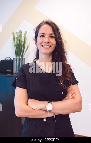Positive Sanitäterin in schwarzer Uniform mit welligem Haar, die in die Kamera schaut und lächelt, während sie in einer modernen Klinik mit gekreuzten Armen steht Stockfoto