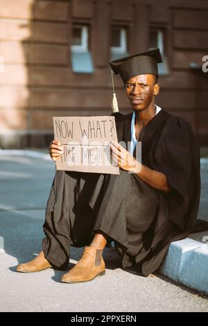 Armer Schwarzer, der auf der Straße mit einem Pappposter auf der Suche nach einem Job sitzt. Universitäts- oder Universitätsstudenten mit Graduiertenkleid und -Mütze. Studieren Sie und Stockfoto