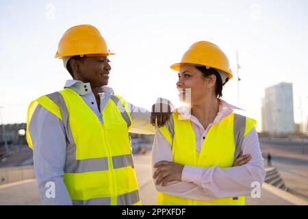 Selbstbewusste afroamerikanische Ingenieurinnen in Schutzhelm und Weste sehen sich gegenseitig an, während sie auf der City Street stehen Stockfoto