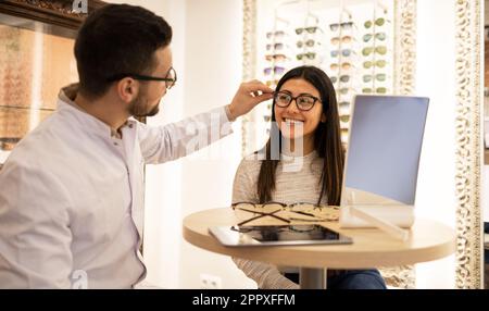 Männlicher Optiker, der lächelnde junge Kundin beim Ausprobieren verschiedener Brillen am Tisch im Optikerladen hilft Stockfoto