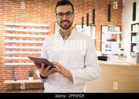Porträt eines lächelnden jungen männlichen Optikers in weißer Uniform und Brille am digitalen Tisch beim Blick in die Kamera im Optikerladen Stockfoto