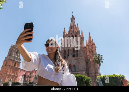 Ein kleiner Winkel glücklicher junger Touristinnen, die Selbstporträts auf dem Handy machen und dabei Zeit während des Ausflugs gegen die antike Kirche in San Miguel de A genießen Stockfoto