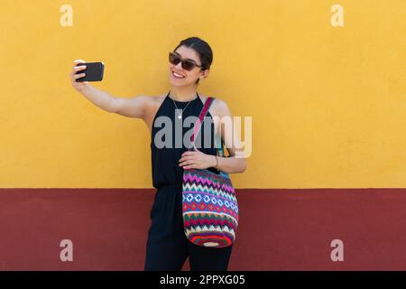 Lächelnde junge Frau in trendigen Outfits und Sonnenbrille mit Tasche, die Selbstfotos auf dem Smartphone macht, während sie in San Miguel de Alle an der gelben Wand steht Stockfoto