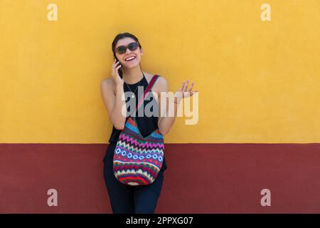 Eine positive junge Dame in trendigen Outfits und einer Sonnenbrille, die lächelt und wegschaut, während sie sich an die gelbe Wand stellt und draußen mit ihrem Smartphone spricht Stockfoto