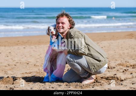 Ein freundlicher Transgender-Mann, nachdem er sich in einen süßen weißen Clumber Spaniel-Welpen verwandelt hat, der in eine transgender-stolze Flagge gewickelt ist und sich am Sandstrand auf s entspannt hat Stockfoto