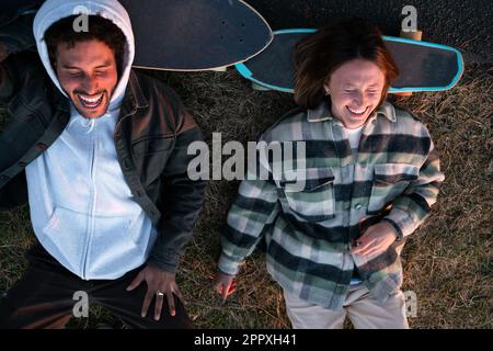 Von oben ein junges, multiethnisches Paar in lässiger Kleidung und mit Skateboards, die auf dem Rasen liegen und mit geschlossenen Augen glücklich lachen, während sie sich entspannen Stockfoto