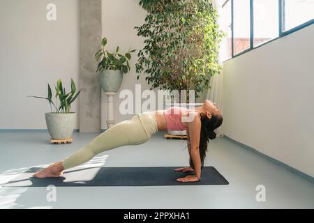 Ganzkörper-Seitenansicht einer fit jungen barfuß Frau in Aktivbekleidung, die Purvottanasana auf Yogamatte aufführt und Achtsamkeit in einem geräumigen Studio übt Stockfoto