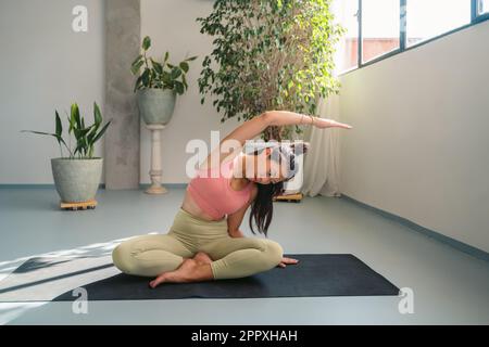 Ein ganzer Körper ruhiger, junger, barfüßiger asiatischer Frau in Sportbekleidung, die in Padmasana sitzt und sich während der Meditation beiseite beugt und den Körper auf y streckt Stockfoto
