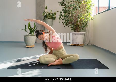 Ein ganzer Körper ruhiger, junger, barfüßiger asiatischer Frau in Sportbekleidung, die in Padmasana sitzt und mit geschlossenen Augen posiert, während sie sich während der Meditation beiseite beugt und str Stockfoto