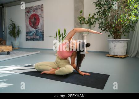 Ein ganzer Körper ruhiger, junger, barfüßiger asiatischer Frau in Sportbekleidung, die in Padmasana sitzt und mit geschlossenen Augen posiert, während sie sich während der Meditation beiseite beugt und str Stockfoto
