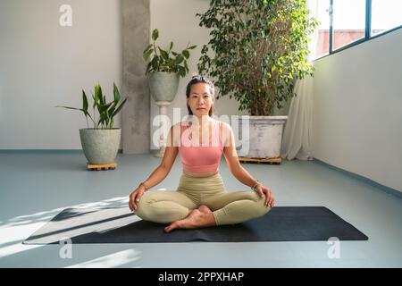 Der ganze Körper einer friedlichen jungen barfüßigen asiatischen Frau in Aktivkleidung meditiert in Padmasana und posiert auf Yogamatte, während sie in modernem Lichtstu in die Kamera schaut Stockfoto