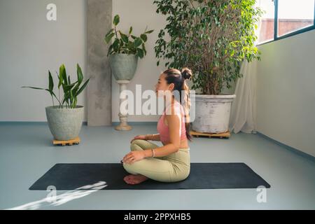 Der ganze Körper einer friedlichen jungen, barfüßigen asiatischen Frau in Aktivbekleidung meditiert in Padmasana und posiert auf Yogamatte, während sie in einem modernen, hellen Studio wegblickt Stockfoto