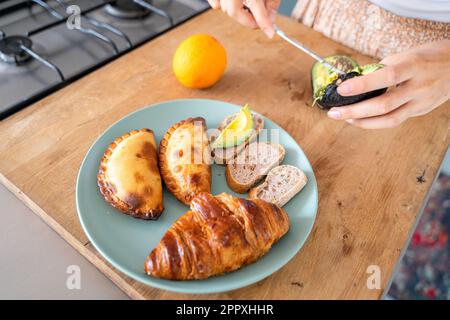 Hoher Winkel des Ernteguts, anonyme weibliche, schälende Avocado mit Messer auf dem Schneidebrett in der Nähe von Orange und Teller mit Empanadas und Croissant während der Zubereitung des Toas Stockfoto
