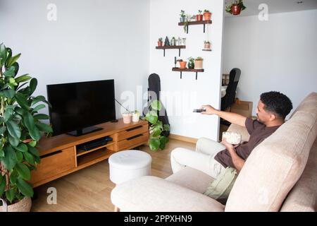 Ein junger afroamerikanischer Mann in legerer Kleidung, sitzt mit einer Schüssel Popcorn und schaltet den Fernseher mit schwarzem Bildschirm ein, während er sich ausruht Stockfoto