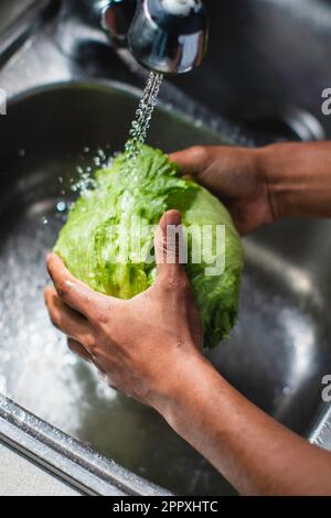 Der große Winkel eines unbekannten afroamerikanischen Chefkochs wäscht frischen grünen Eisbergsalat in einem Metallwaschbecken mit Wasser, während er gesunden Salat in der Küche kocht Stockfoto