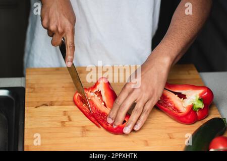 Konzentrierter, nicht wiedererkennbarer afroamerikanischer Mann in weißem T-Shirt, schneidet reife rote Paprika mit scharfem Messer auf Holzhacke, während Präpar Stockfoto