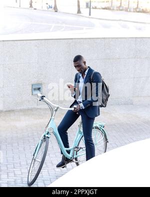 Seriöser afroamerikanischer Geschäftsmann in einem formellen Anzug, der auf dem Fahrrad sitzt und auf dem Handy surft, während er auf der Straße in der Nähe einer Betonwand fährt Stockfoto