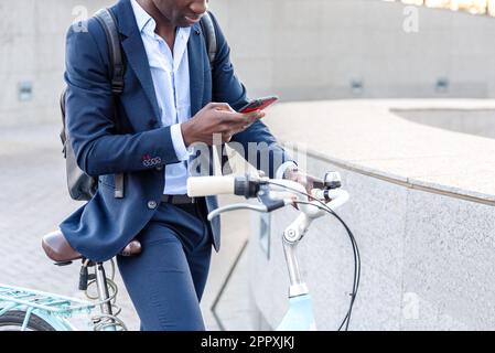 Gekürzter, nicht wiedererkennbarer afroamerikanischer Geschäftsmann in einem formellen Anzug, sitzt auf dem Fahrrad und surft auf dem Handy, während er auf der Straße in der Nähe von Concr fuhr Stockfoto