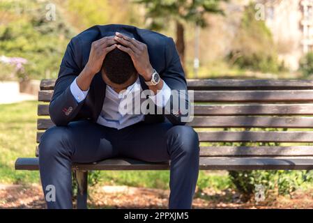 Ein afroamerikanischer Geschäftsmann im formellen Anzug, der mit den Händen am Kopf sitzt, während er nach dem Arbeitstag Probleme vor verschwommenem Hintergrund hat Stockfoto