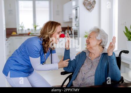 Eine Schwester mit Clownnnase, die Spaß mit ihrer Seniorin-Klientin hat. Stockfoto