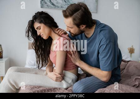 Bärtiger Mann beruhigt enttäuschte Freundin im Pyjama im Schlafzimmer, Stock Image Stockfoto