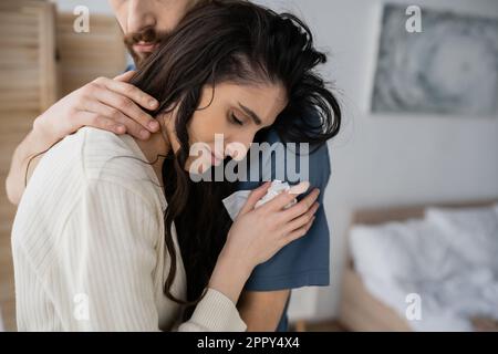Ein bärtiger Mann beruhigt die traurige Freundin mit einer Serviette im verschwommenen Schlafzimmer, ein Bild der Aktie Stockfoto