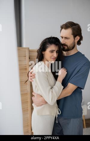 Ein bärtiger Mann im Pyjama umarmt eine unzufriedene Freundin zu Hause, ein Image Stockfoto