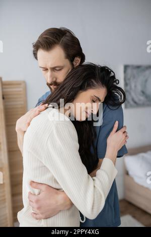 Bärtiger Mann umarmt und beruhigt verärgerte Freundin im Schlafzimmer zu Hause, Stock Image Stockfoto