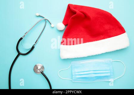 Medizin-weihnachts-Neujahrskonzept während der Coronavirus-Covid-Zeit. Medizinisches Stethoskop und Schutzmaske mit weihnachtsmannmütze auf blauem Hintergrund. Stockfoto