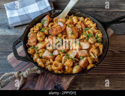 Gebratener Lachs in der Pfanne mit Muschelnudeln und cremiger Tomatensoße in einer gusseisernen Pfanne, isoliert auf einem Holztisch Stockfoto