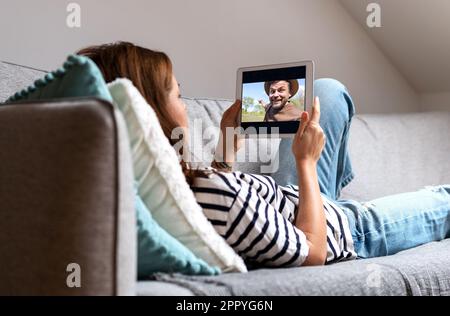 Frau, die zu Hause auf dem Sofa liegt und sich mit ihrem digitalen Tablet im Internet ein Video-Blog ansieht. Stockfoto