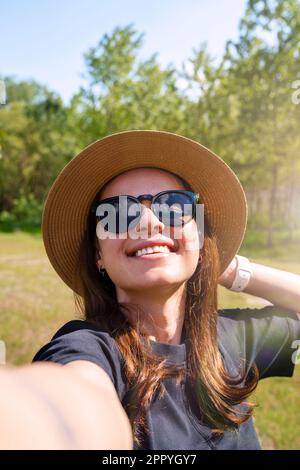 Junge Frau mit Sonnenbrille und Strohhut lacht und macht ein Selfie, während sie im Sommer im Freien spaziert. Stockfoto