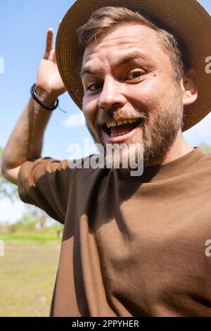 Ein junger erwachsener Mann mit Strohhut lächelt und macht ein Selfie, während er draußen spaziert. Stockfoto